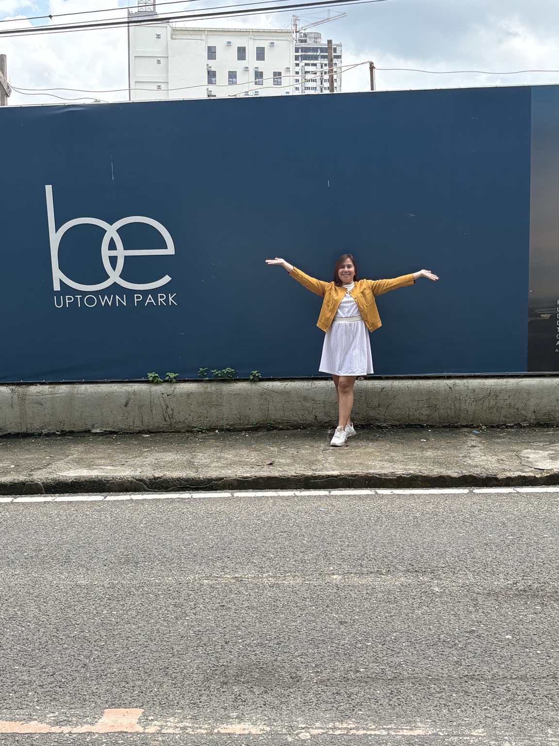 Nessa smiling with open arms beside the BE Uptown Park site sign across from STC